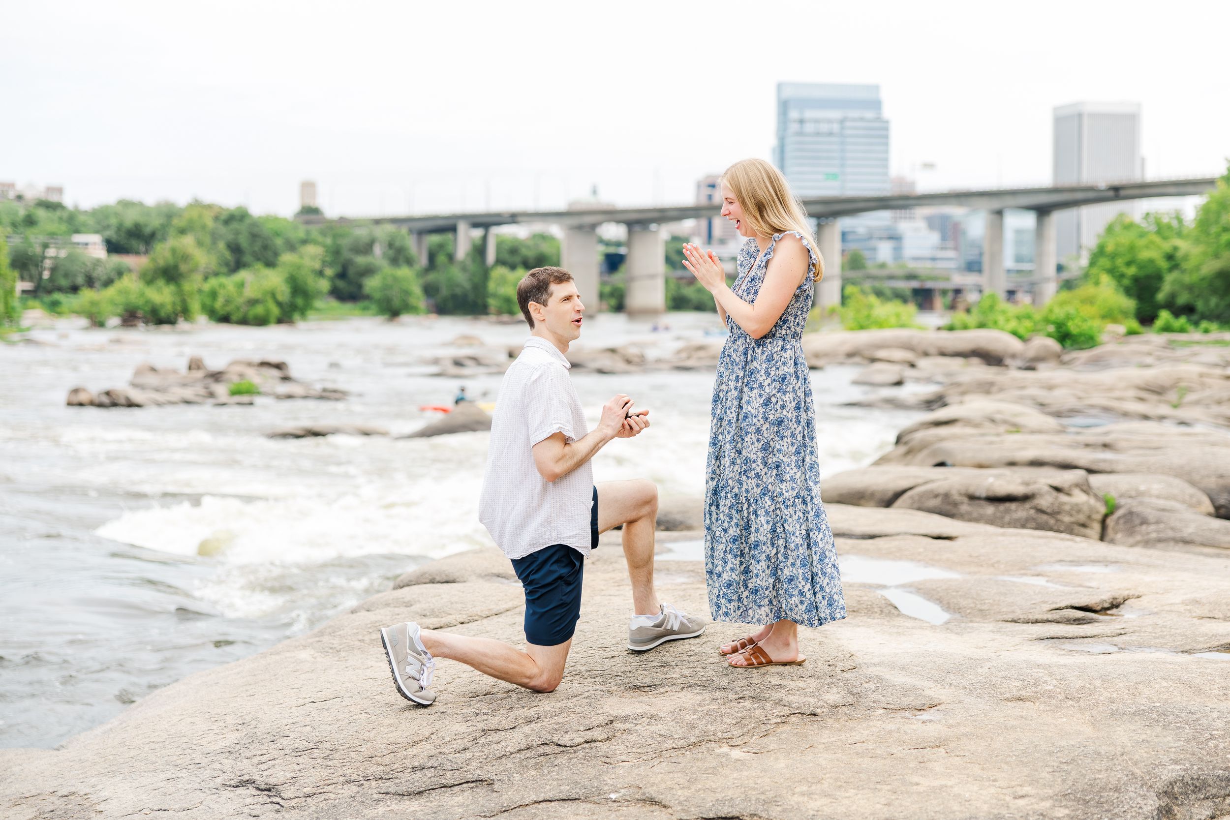 Man proposes on one knee to woman on rocky riverbank with bridge in background.