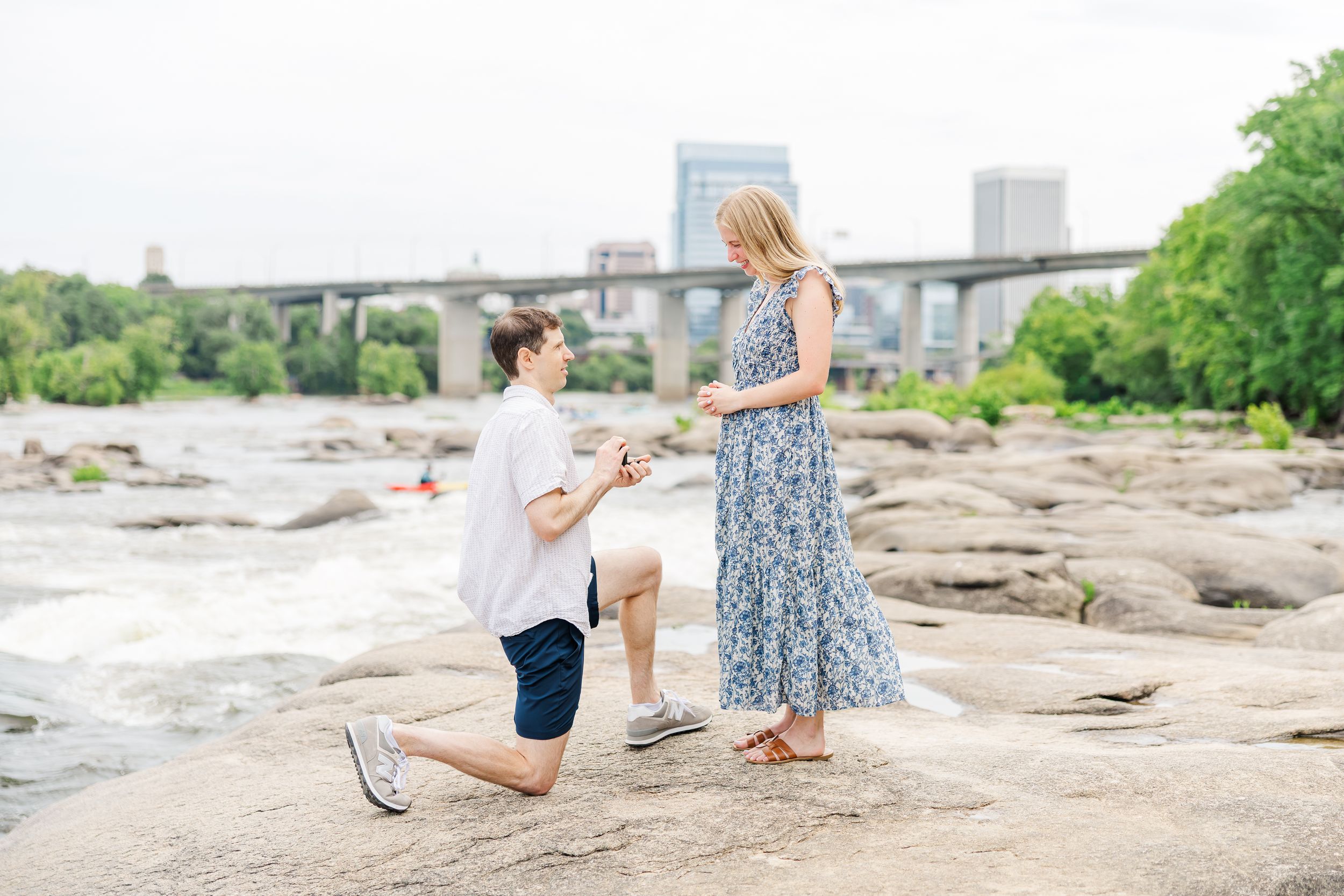 Man kneeling on rocks proposing to woman in floral dress near river with city bridge in background.