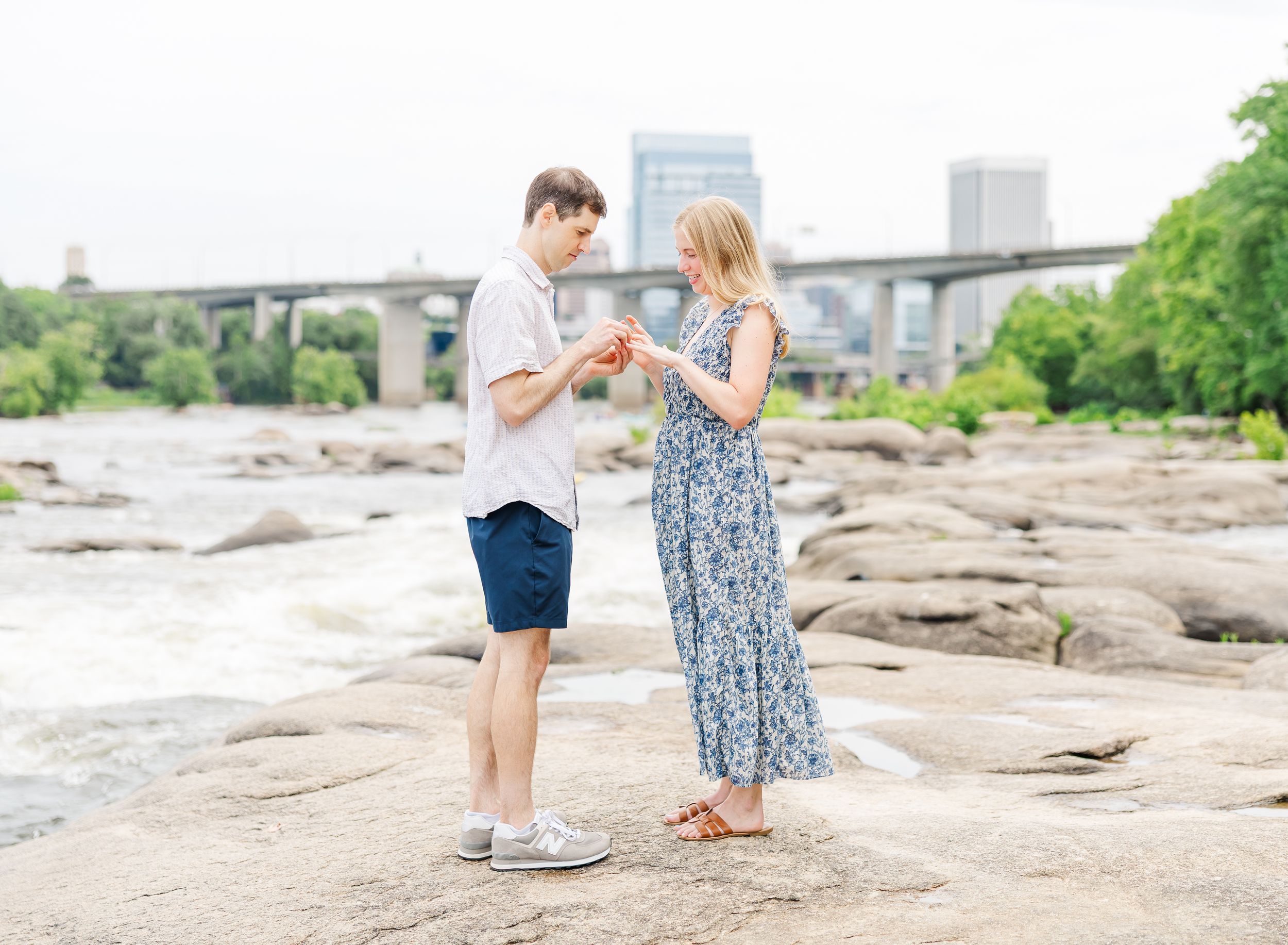 Couple sharing a romantic proposal moment on rocky riverbank with city bridge skyline backdrop.
