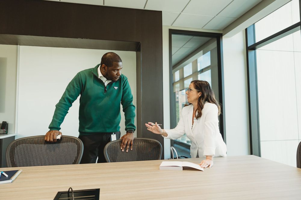 Two colleagues discussing work in a modern conference room, man in green hoodie and woman in white blouse.
