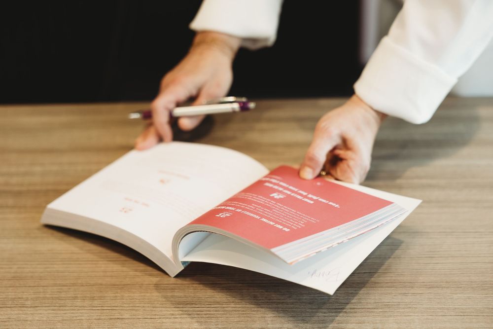 Person in white shirt reviewing an open book with a red page on a wooden desk, holding a pen.