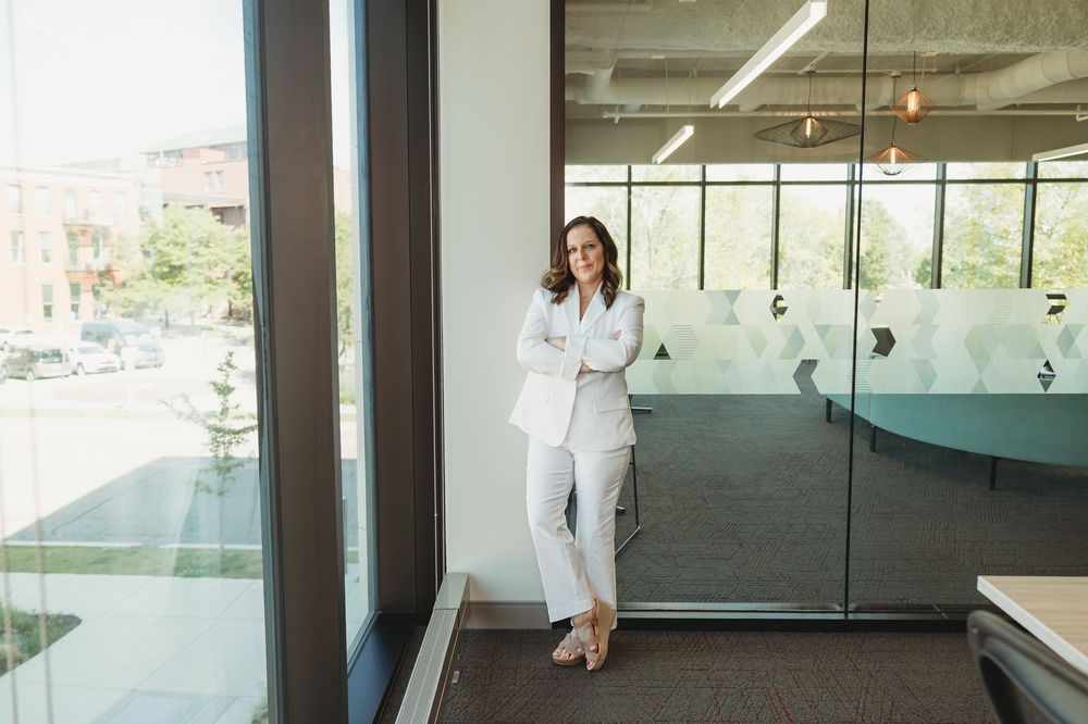 Professional woman in white suit leaning against glass office wall, smiling confidently in modern workspace.