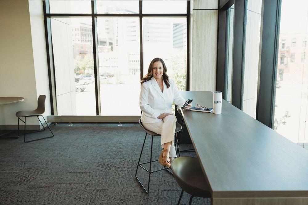 Professional woman in white blazer sitting confidently at a modern office conference table near large windows.