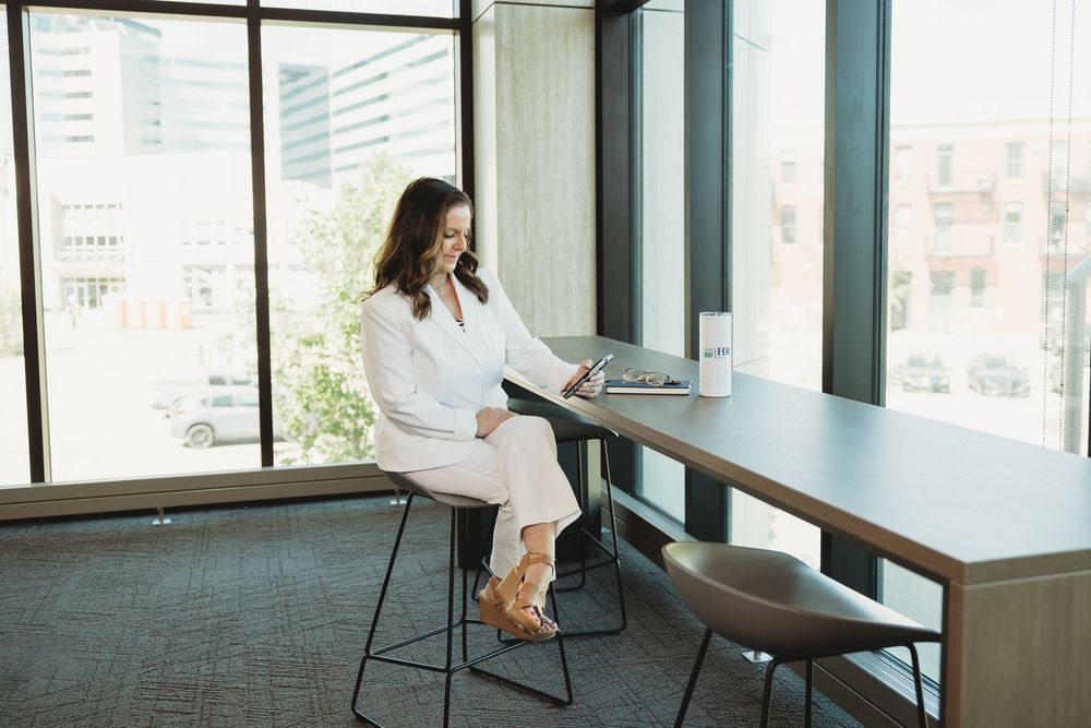 Professional woman in white outfit sitting on barstool using smartphone in modern office lobby.