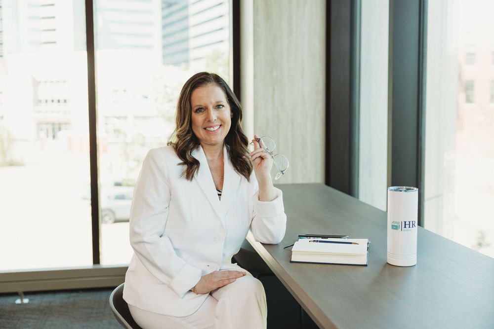 Professional woman in white blazer smiling at desk with notebook and water bottle in modern office.