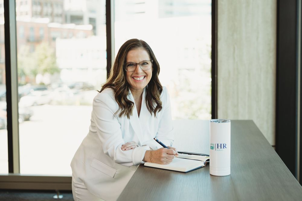 Professional woman in white blazer smiling at desk with notebook and branded tumbler in modern office.
