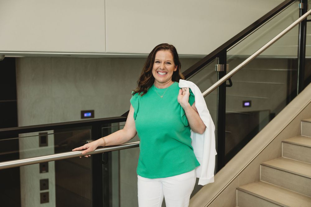 Woman in green top and white pants smiling on modern office staircase with glass railing.