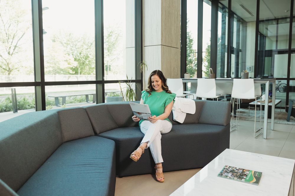 Woman in green top sitting on gray sectional sofa using laptop in modern bright office lobby.