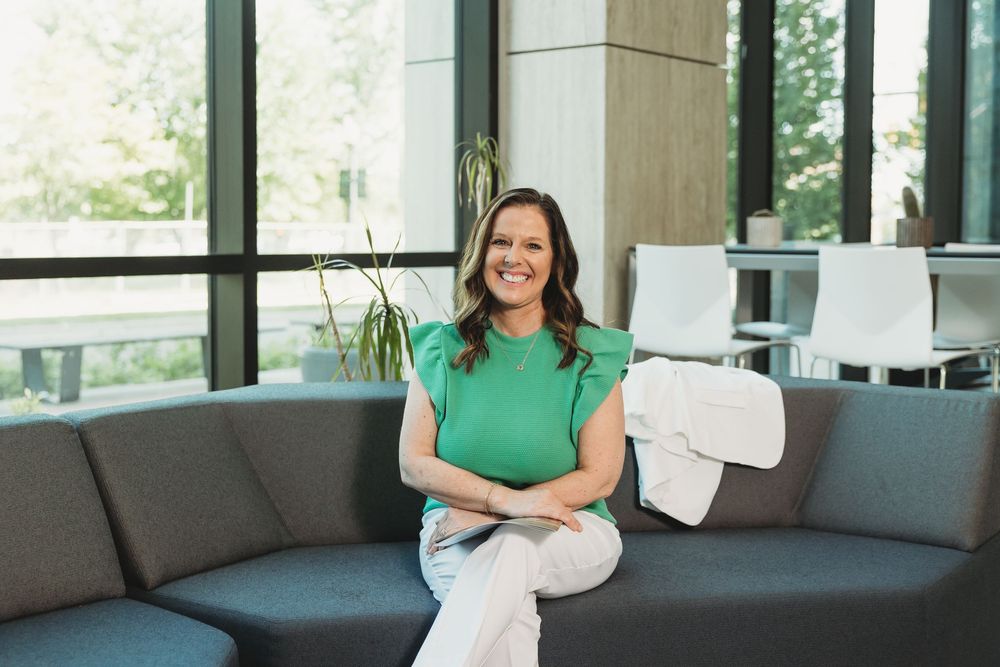 Smiling woman in green top sitting on gray sofa in modern office lobby with large windows.