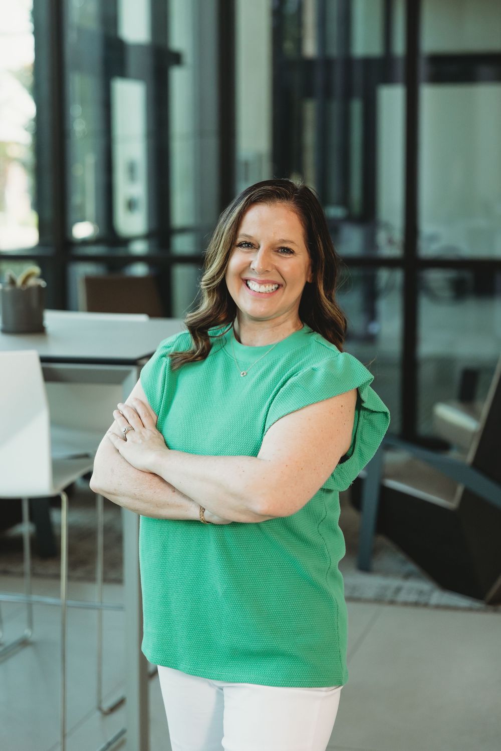 Smiling woman in green top with arms crossed standing in modern office lobby.