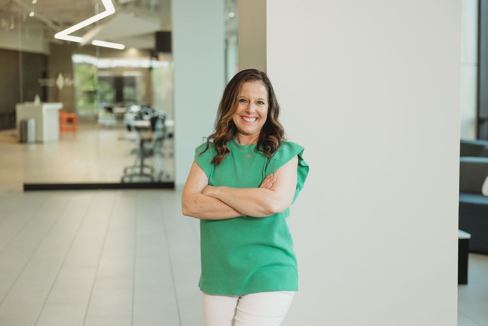 Professional woman in green top smiling with arms crossed in modern office lobby.