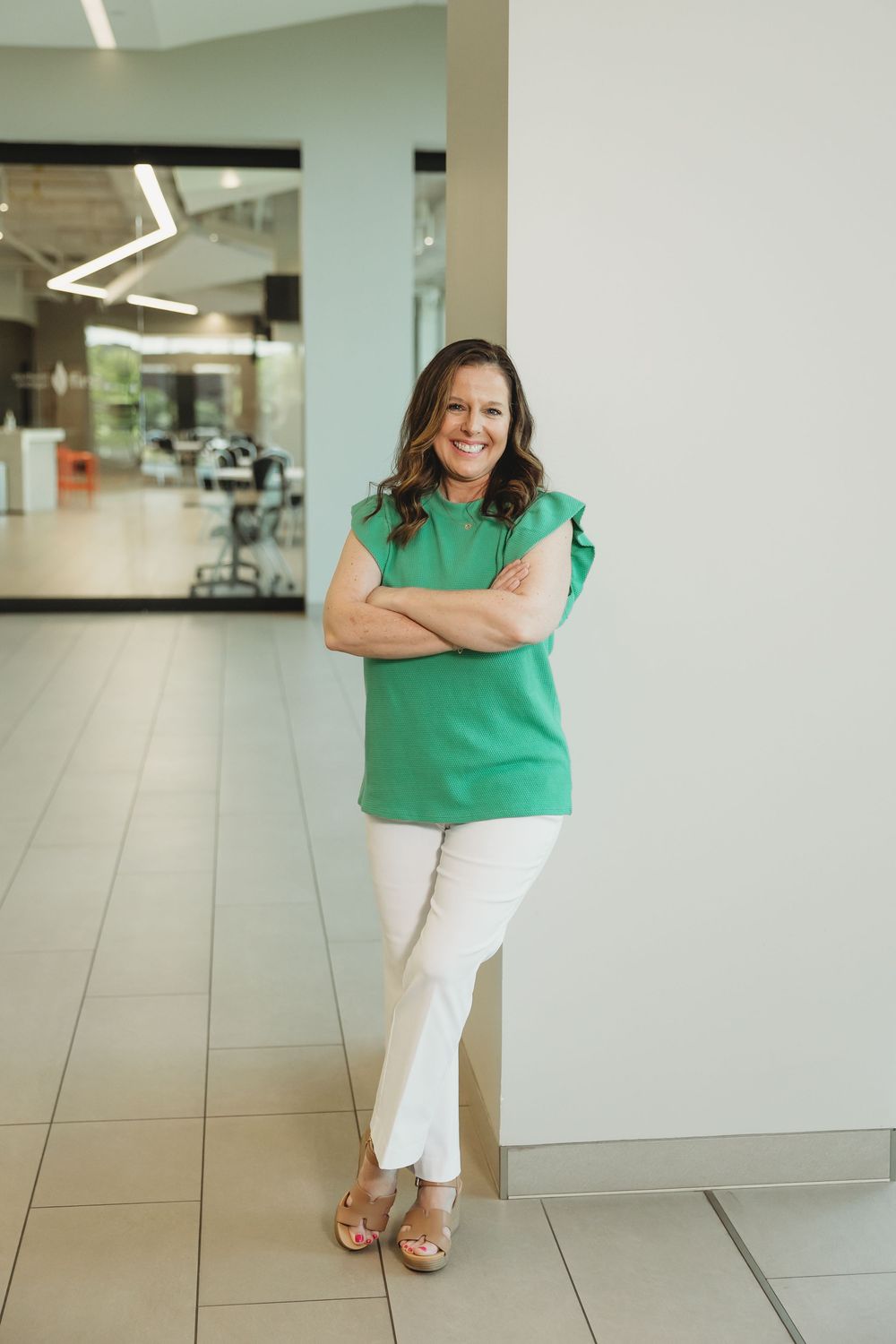 Woman in green top and white pants smiling with arms crossed, leaning against wall in modern office.