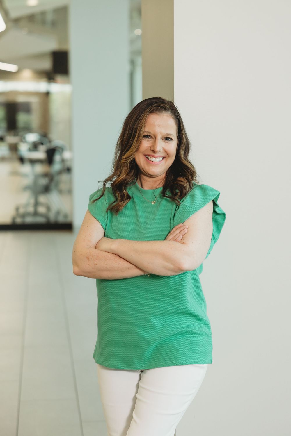 Professional woman in green top smiling with arms crossed in modern office hallway.