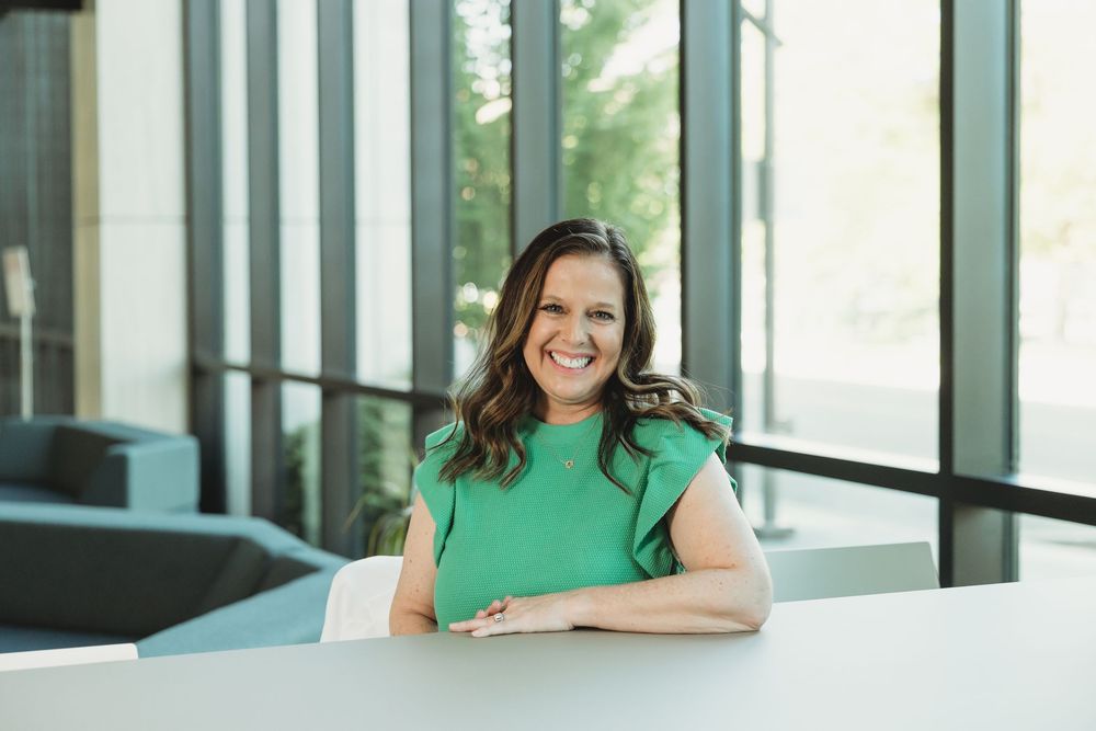 Woman in green top smiling at white table in modern office with large windows.