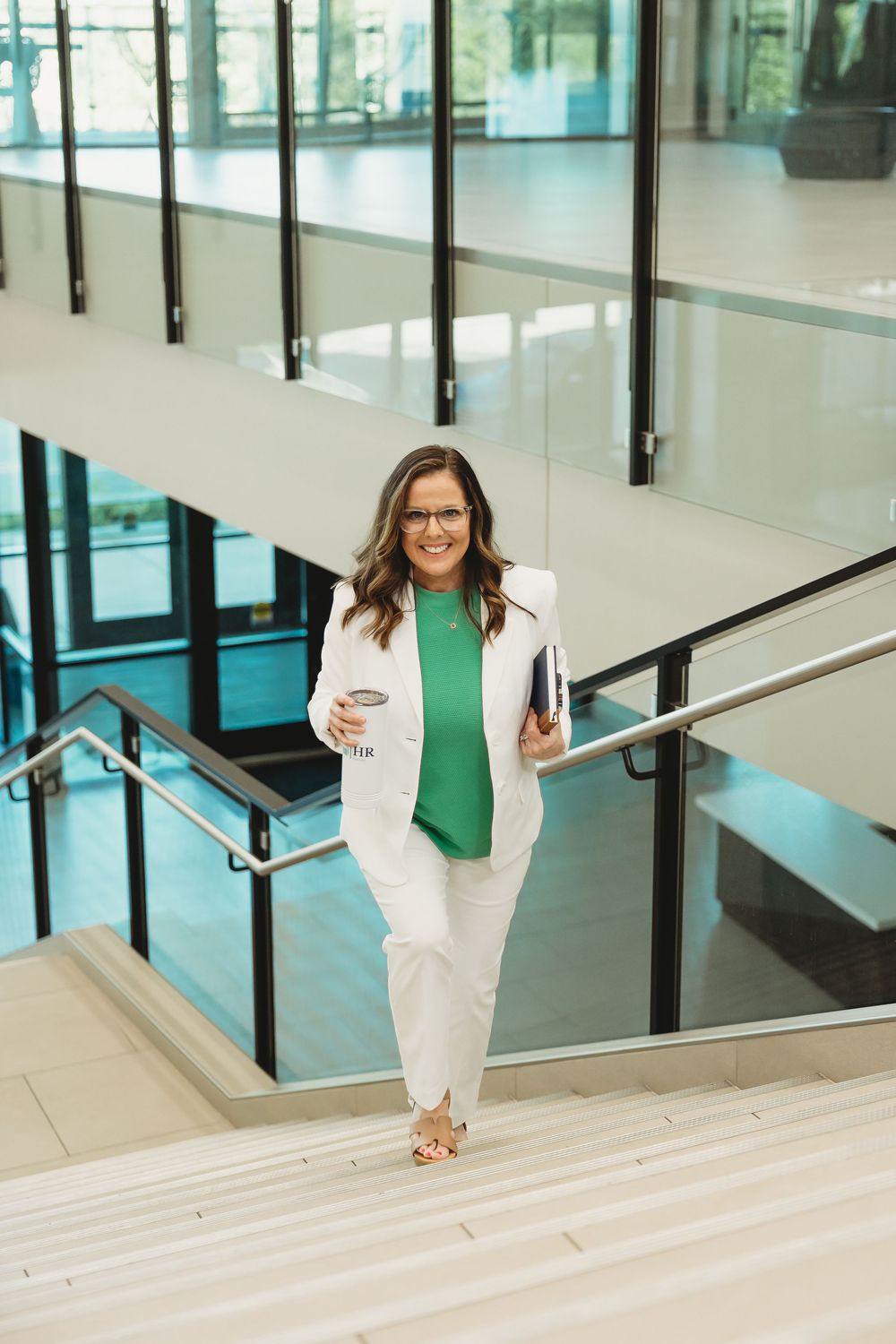 Professional woman in white suit and green top walking down modern office stairs holding coffee and notebook.