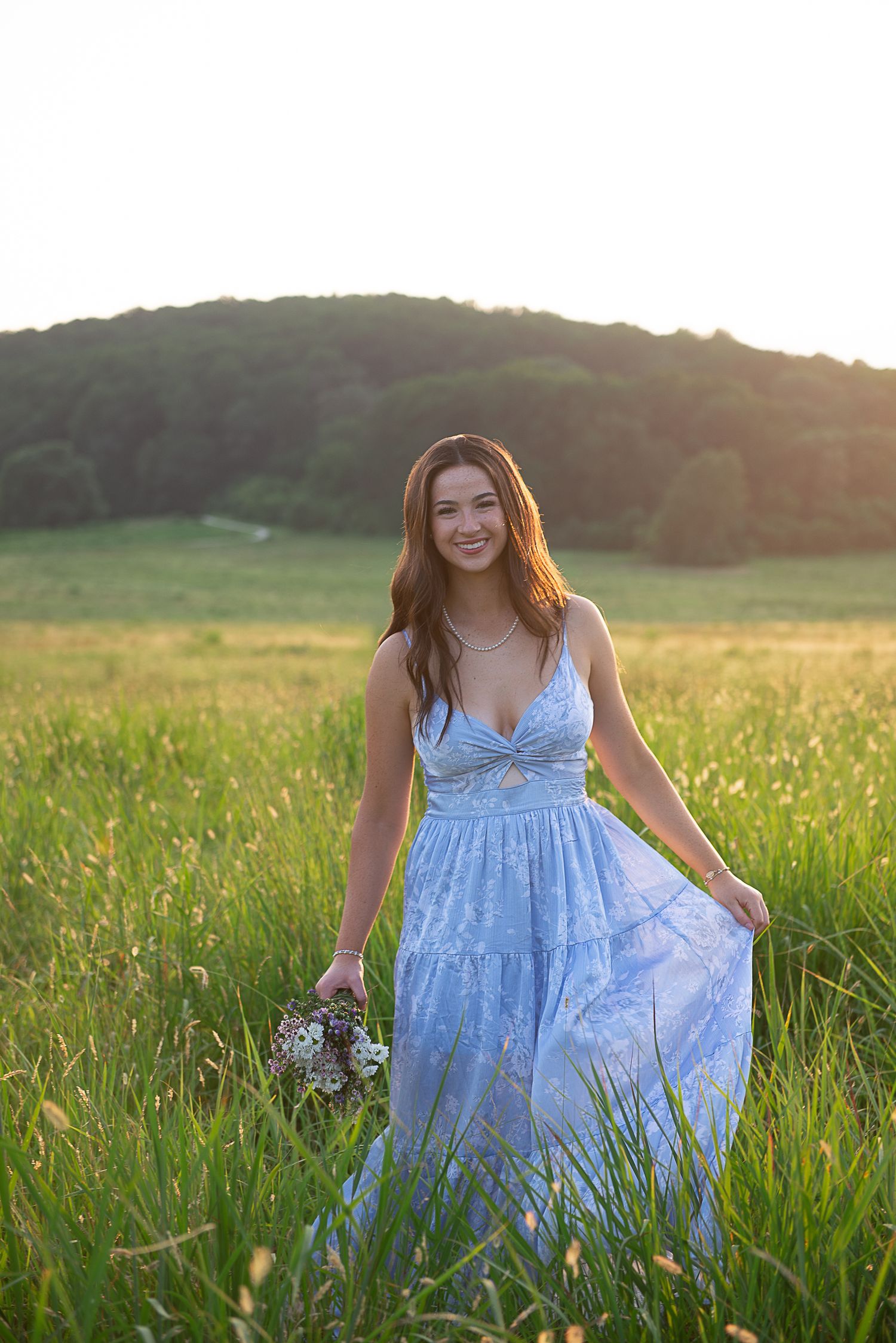 Young woman in blue floral dress holding wildflowers in a sunlit green meadow with hills in background.