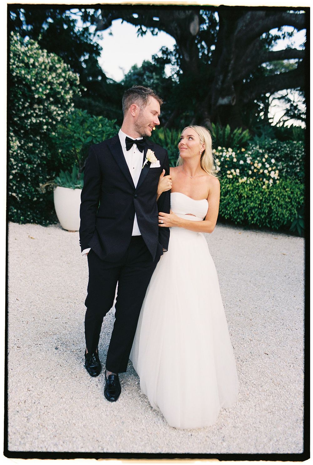 Bride in strapless white gown and groom in black tuxedo share a romantic moment outdoors at their wedding.