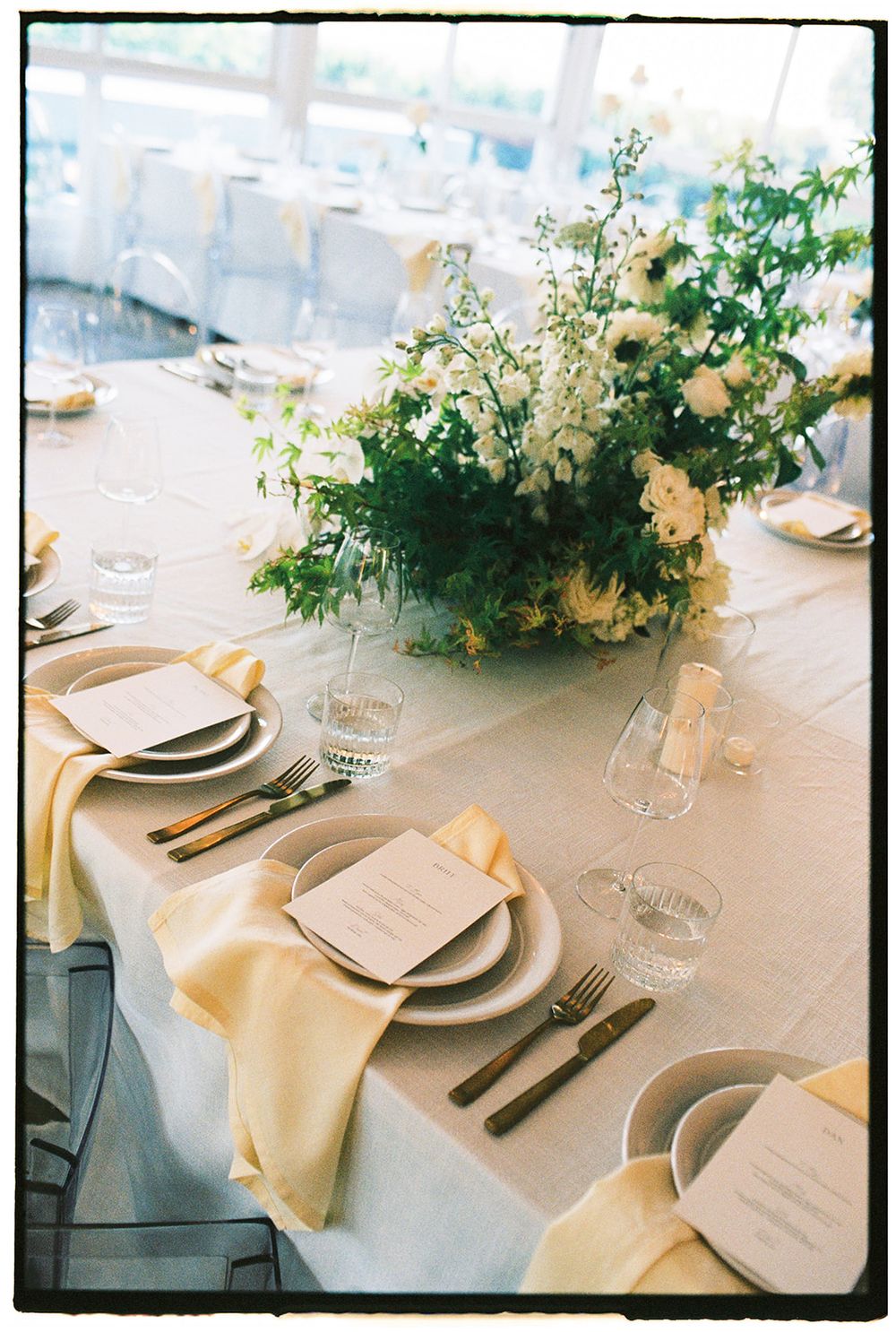Elegant wedding reception table with yellow napkins, white floral centerpiece, crystal glassware, and menu cards on white linen.