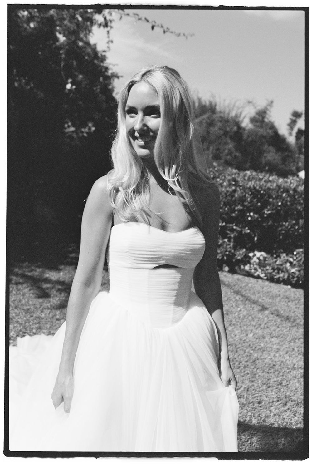 Smiling blonde bride in strapless white wedding gown posing outdoors in garden, black and white photo.