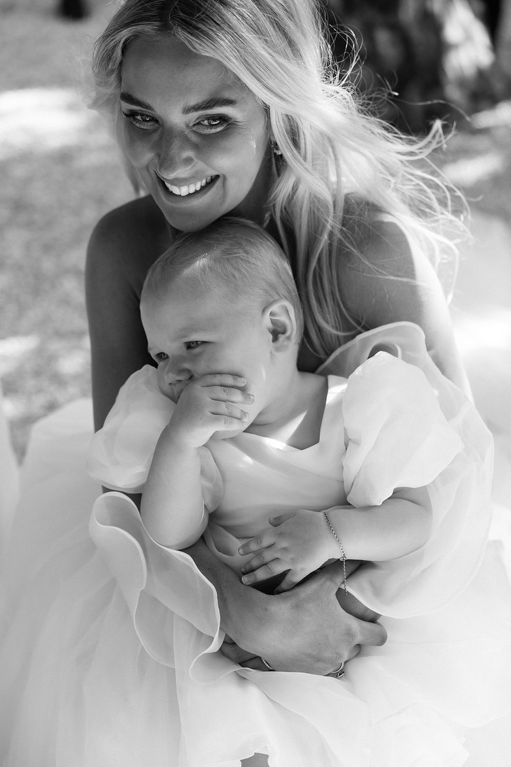 Black and white photo of a smiling blonde woman in a white dress tenderly holding a baby outdoors.