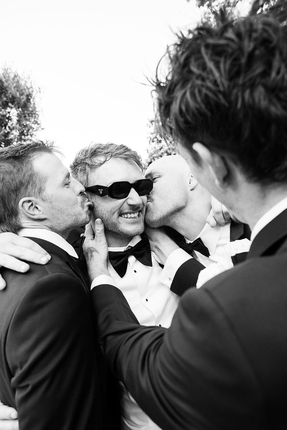Groom in tuxedo and sunglasses laughs as two groomsmen kiss his cheeks at wedding celebration.