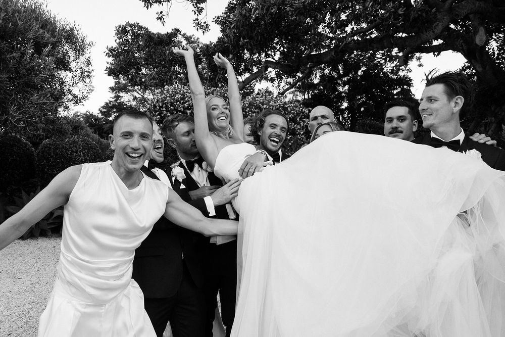 Black and white wedding photo of bride, groom, and bridal party celebrating joyfully outdoors.