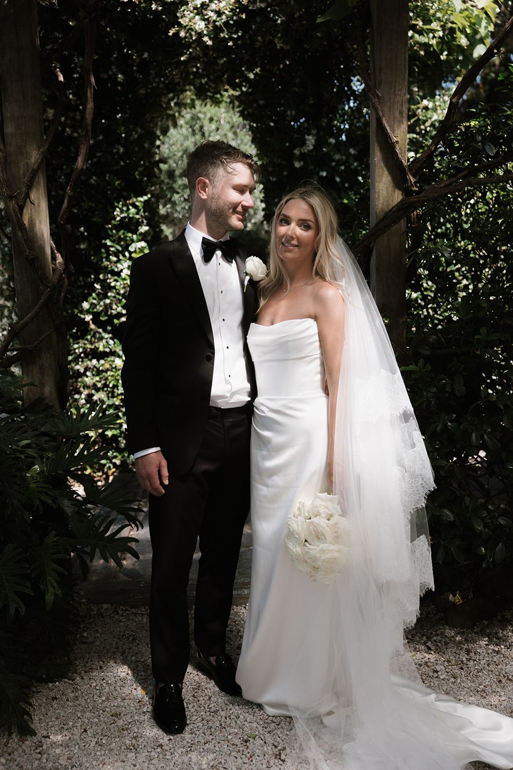 Bride in strapless white gown with veil and groom in black tuxedo pose together in a lush garden setting.