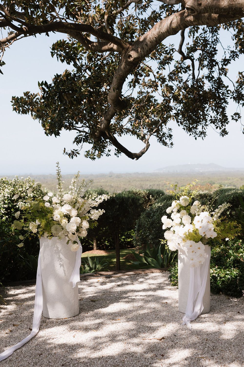 Elegant outdoor wedding ceremony arch with white floral arrangements on pedestals beneath a sprawling oak tree.