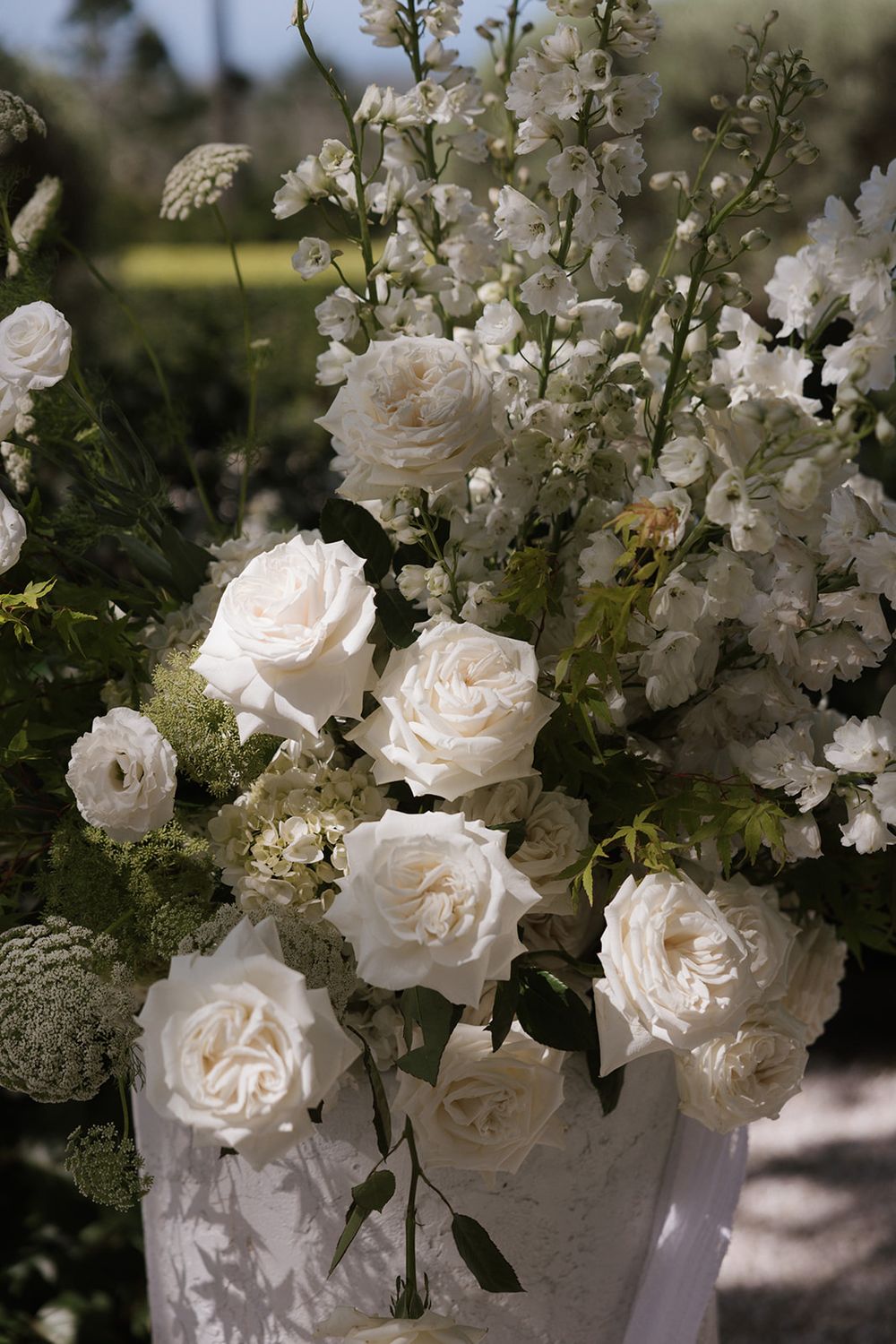 Elegant white rose wedding floral arrangement with lush greenery in a white vase outdoors.