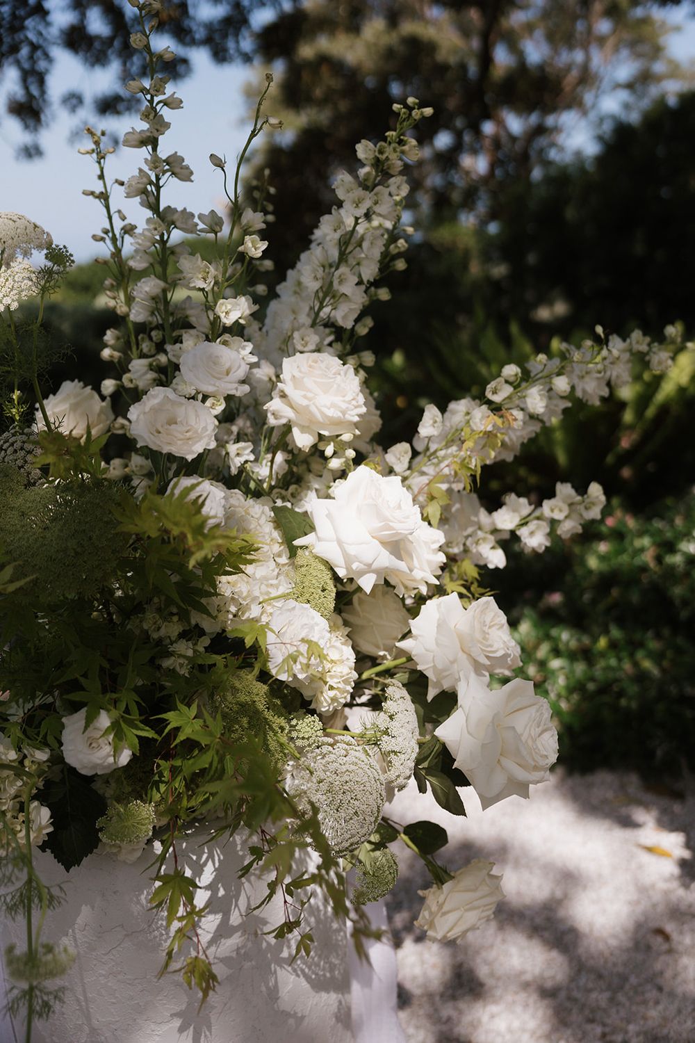 Close-up of lush white wedding floral arrangement featuring roses, hydrangeas, and delphinium in garden setting.