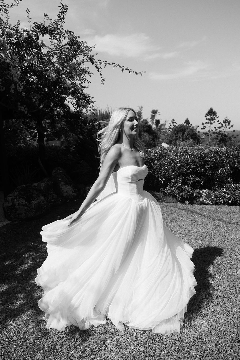 Bride in flowing strapless ball gown twirling in garden, black and white photo.