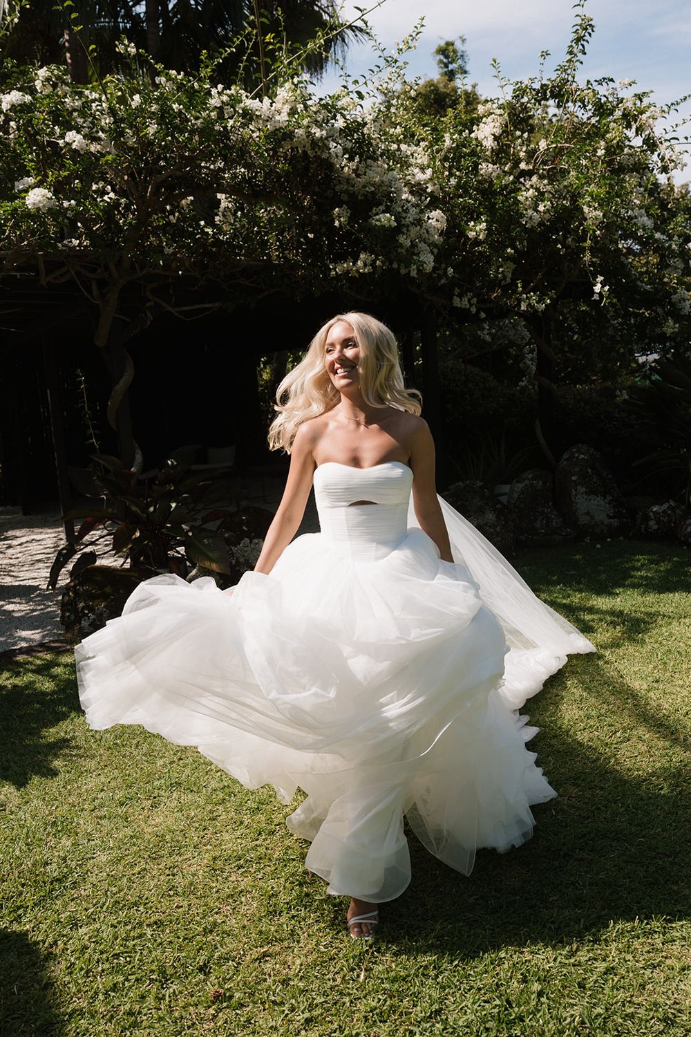 Bride in a strapless white tulle ballgown twirling joyfully in a lush garden on her wedding day.
