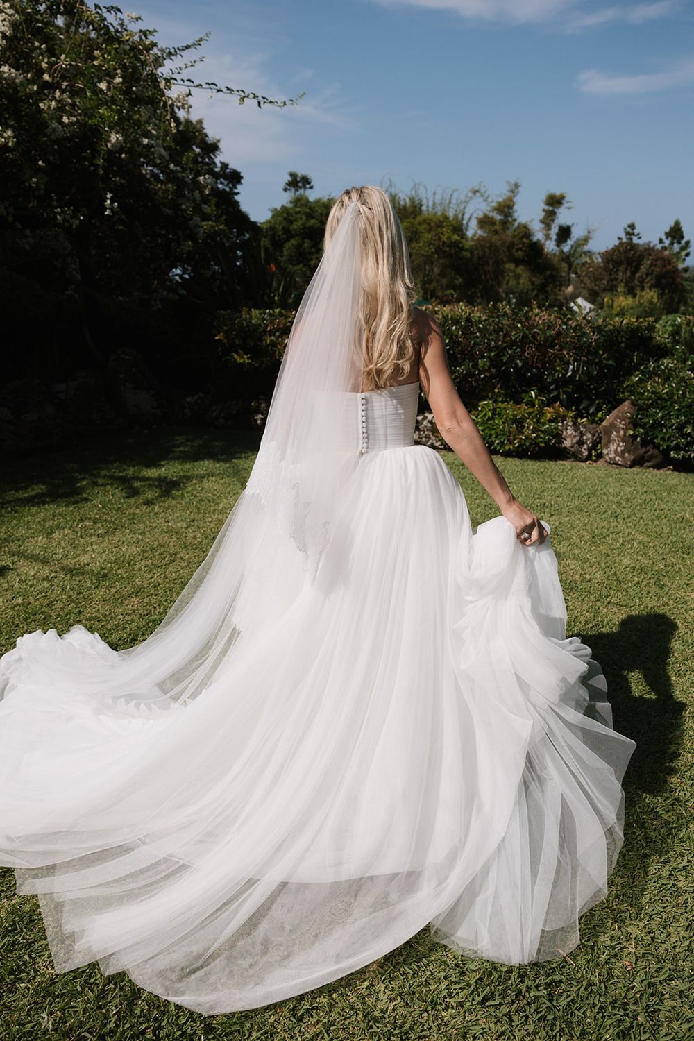 Bride in white tulle ball gown with veil stands in lush garden, viewed from behind, holding dress train.