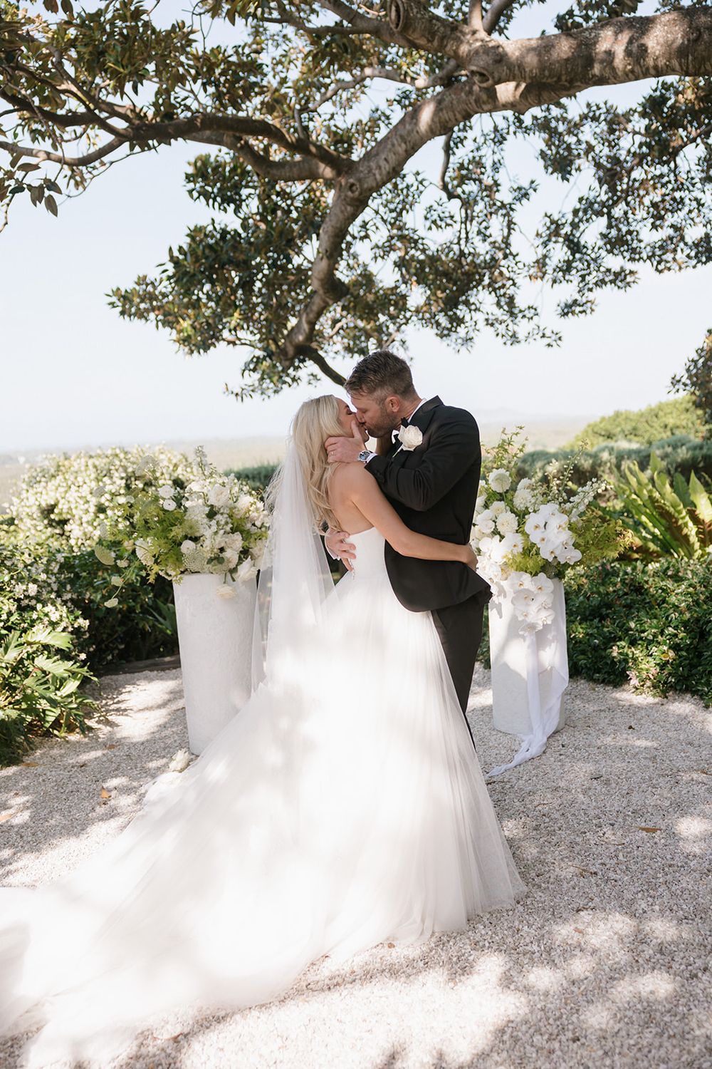 Bride and groom share first kiss under oak tree at outdoor wedding ceremony with white floral arrangements.
