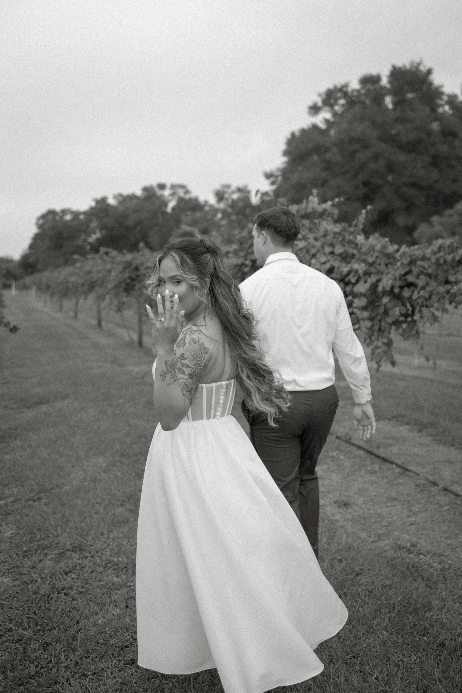 Black and white photo of a bride showing her ring while walking with groom through a vineyard.
