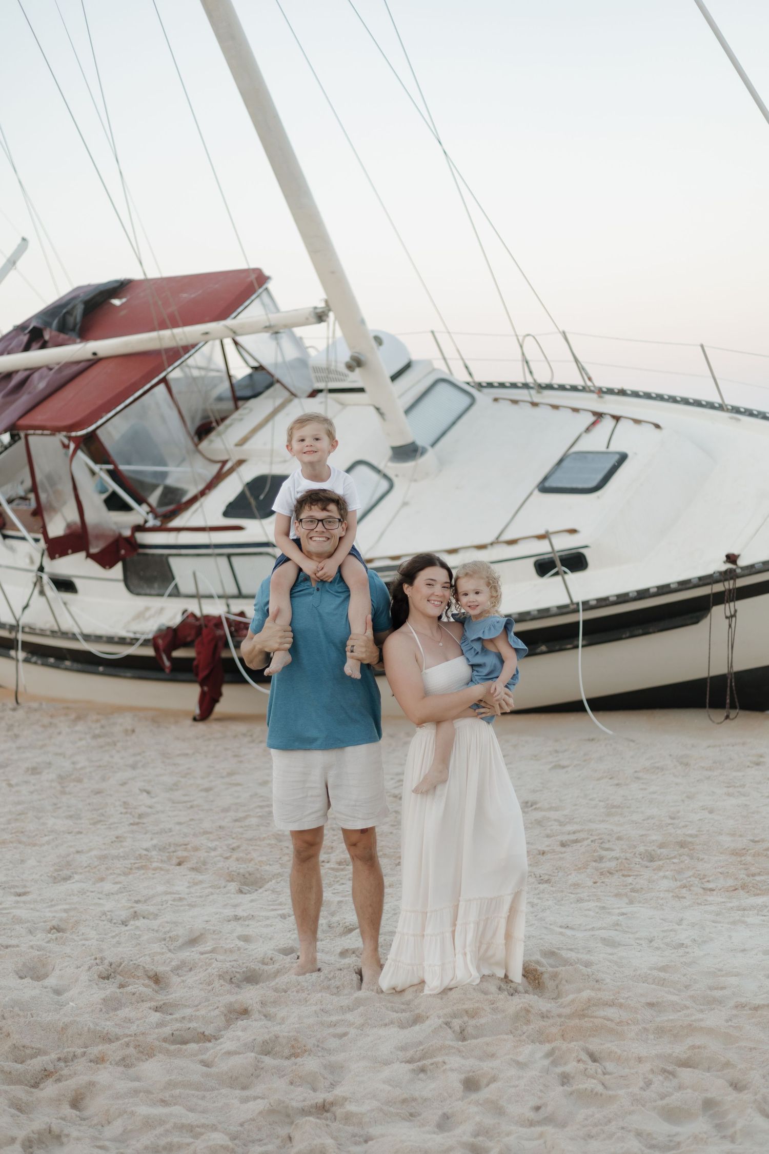 Family of four posing on sandy beach with sailboats, parents holding two young children, wearing casual summer outfits.