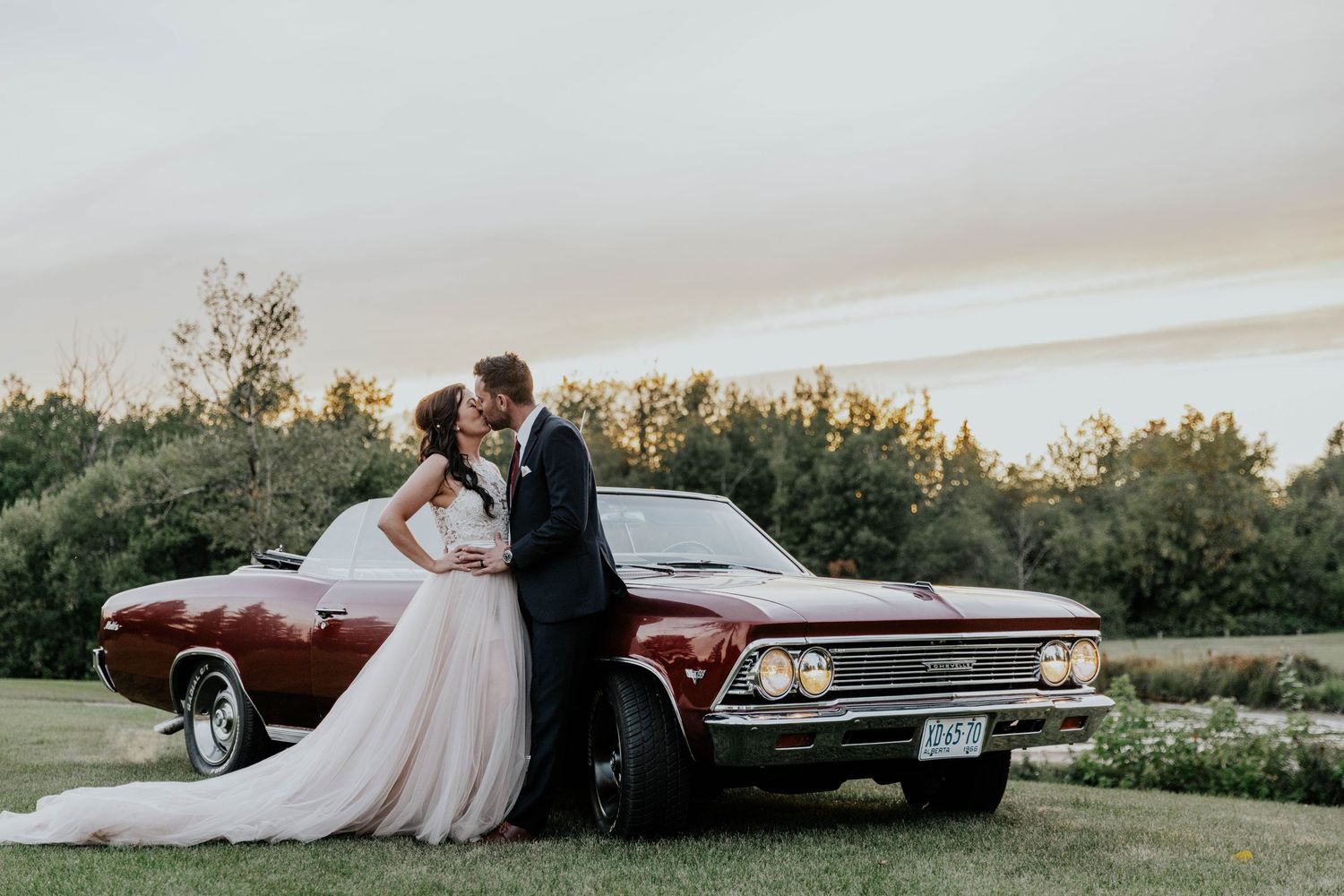 Bride and groom kissing beside a classic red convertible car at sunset on their wedding day.