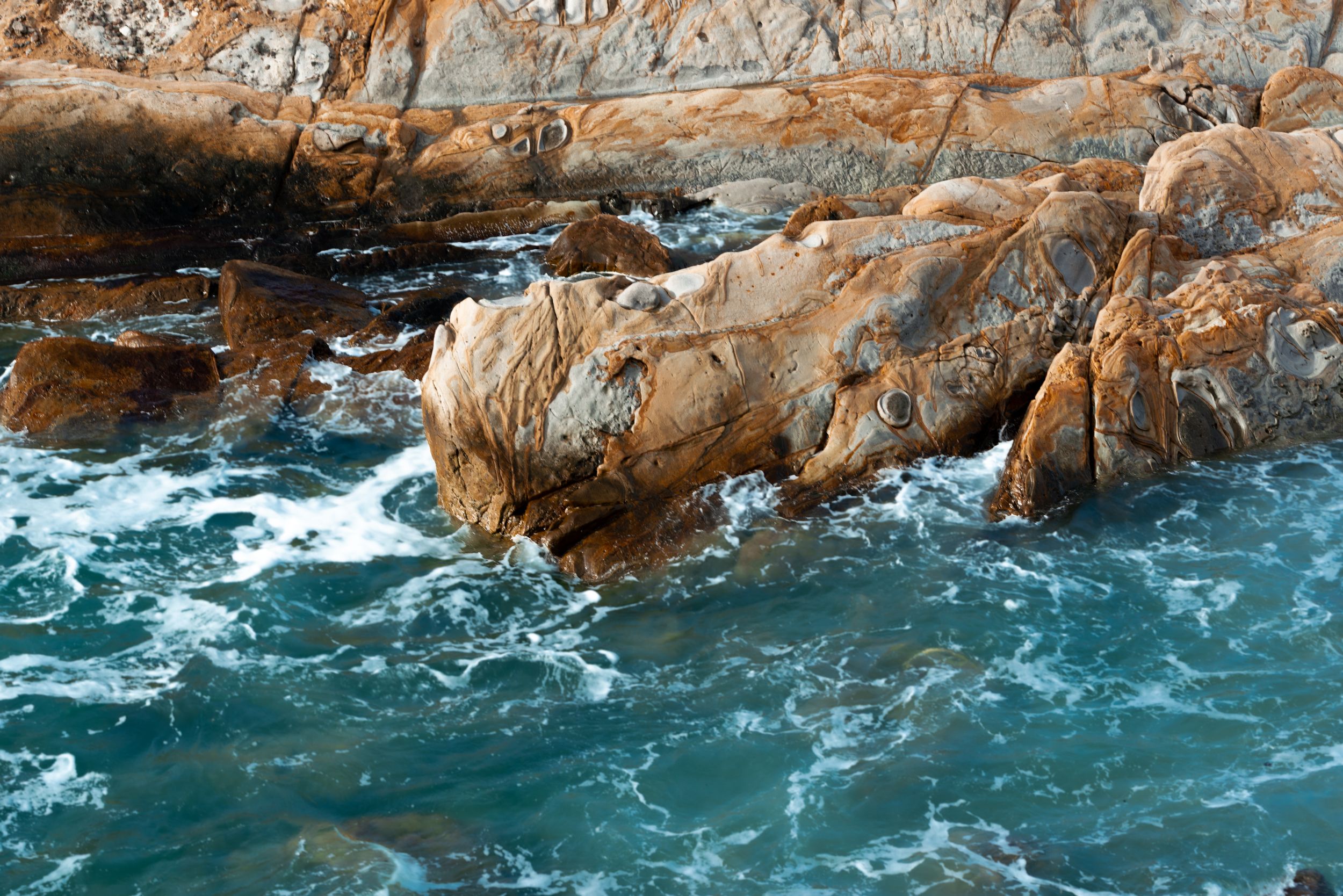 Rocky coastal shoreline with turquoise waves crashing against orange-brown rocks along a rugged ocean coast.
