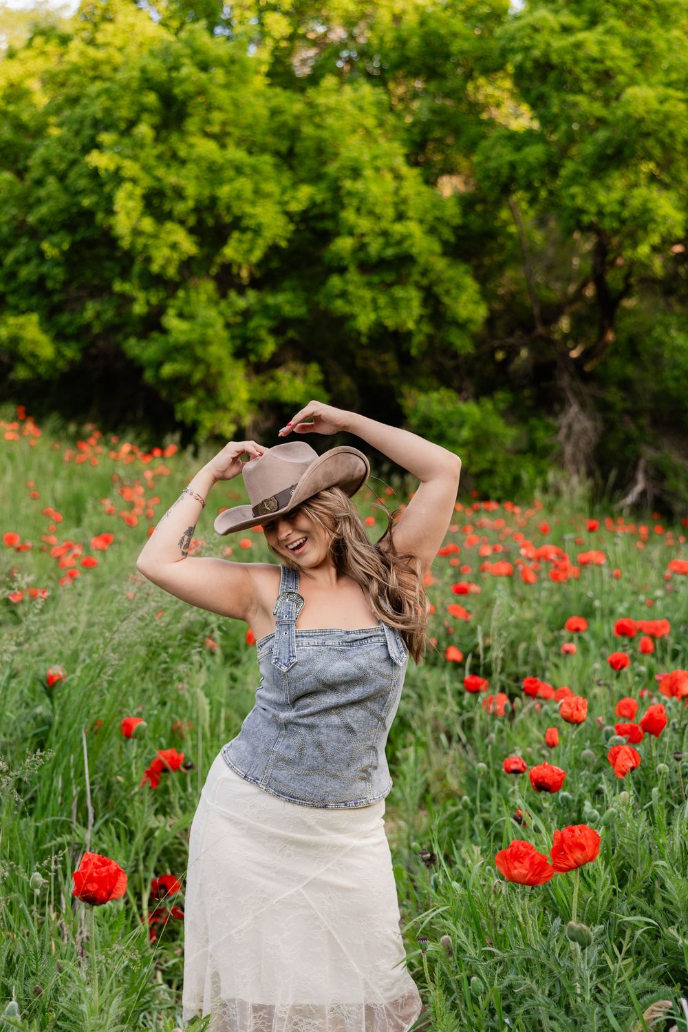 Woman in denim overalls and cowboy hat poses in a vibrant red poppy field with lush green trees.