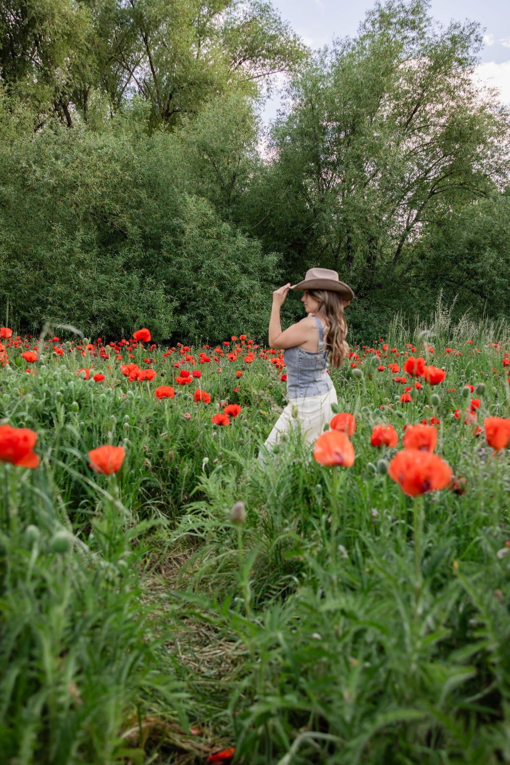 Woman in hat and white skirt walking through vibrant red poppy field surrounded by lush green trees.