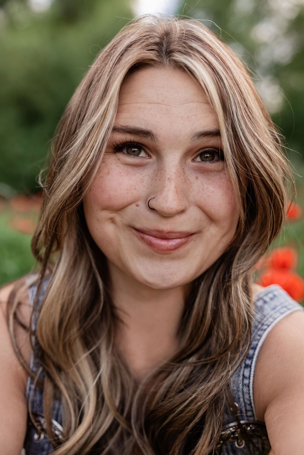 Young woman with freckles, highlighted hair, and nose ring smiling outdoors near colorful flowers.