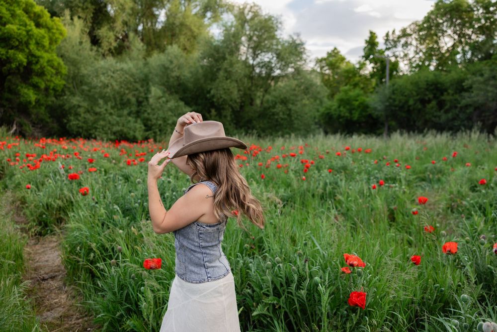 Woman in cowboy hat and white skirt standing in a vibrant red poppy field surrounded by lush green trees.