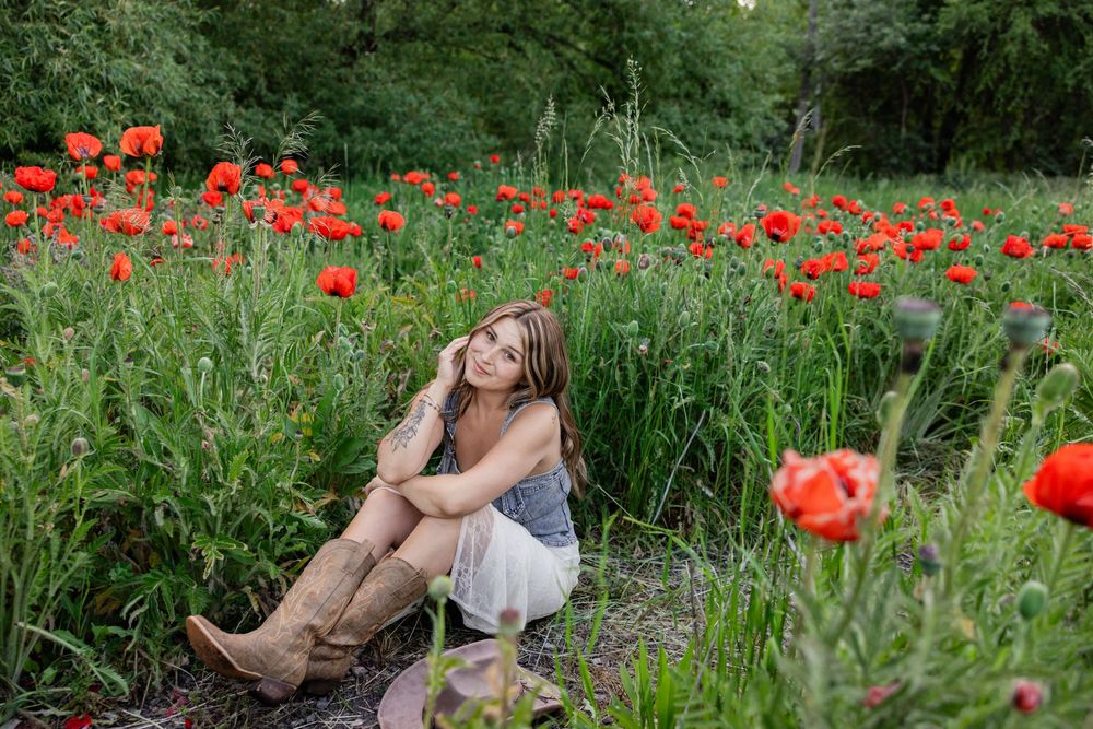 Young woman in cowboy boots sits among vibrant red poppy flowers in a lush green field, gazing upward dreamily.