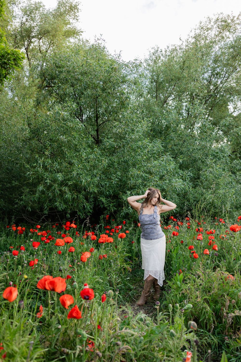 Woman in gray top and white skirt standing in a vibrant red poppy field surrounded by lush green trees.