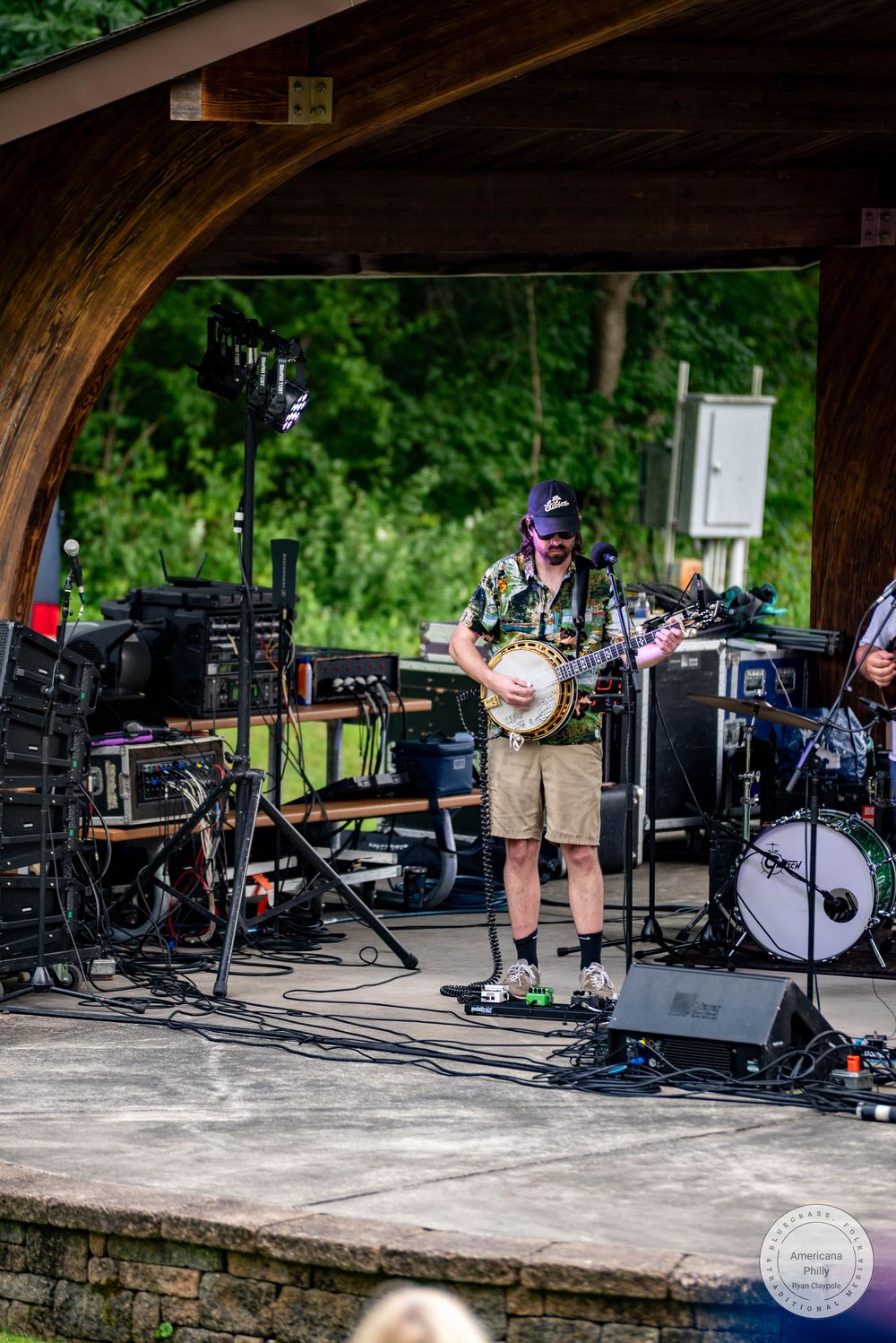Musician playing banjo on outdoor stage at park pavilion with band equipment and lush green trees in background.