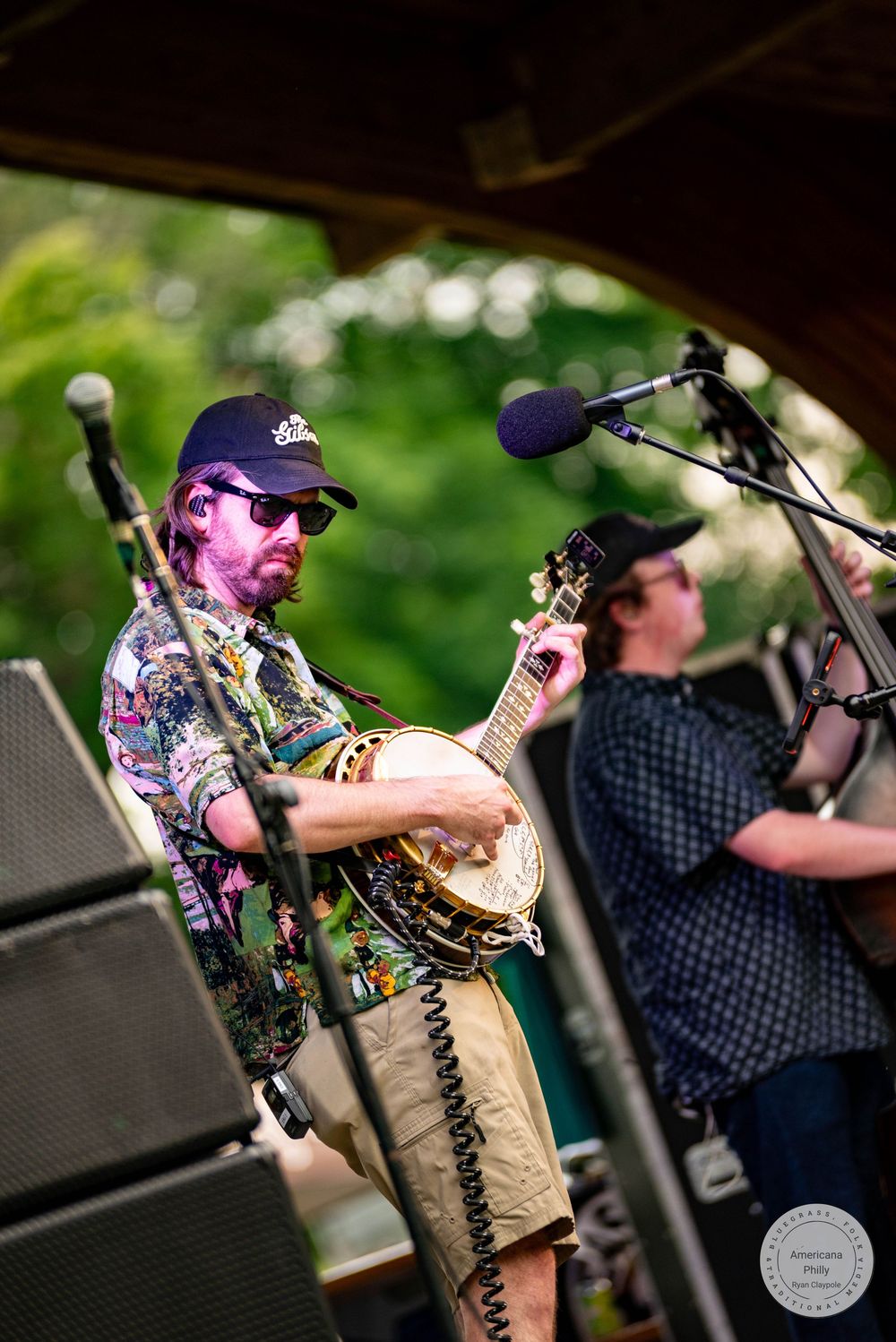 Musician playing banjo on outdoor stage wearing floral shirt, cap, and sunglasses at a live music festival.