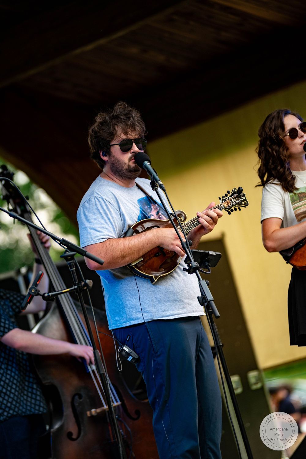 Man playing mandolin on outdoor stage with band members including bassist and guitarist at live music festival.