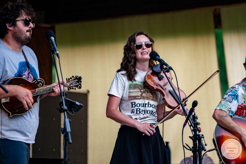Woman in Bourbon & Beyond shirt plays violin on outdoor stage with band members playing mandolin nearby.