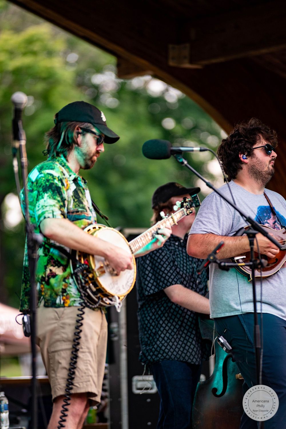 Three musicians perform on outdoor stage, featuring banjo, mandolin, and guitar in colorful festival setting.