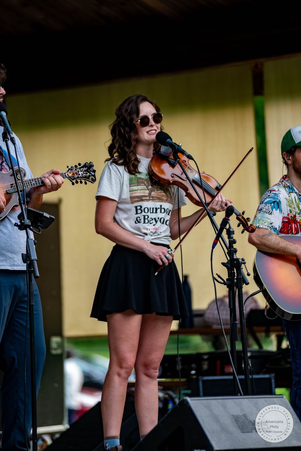 Woman in Bourbon & Beyond shirt plays violin on outdoor stage with band members playing mandolin and guitar.